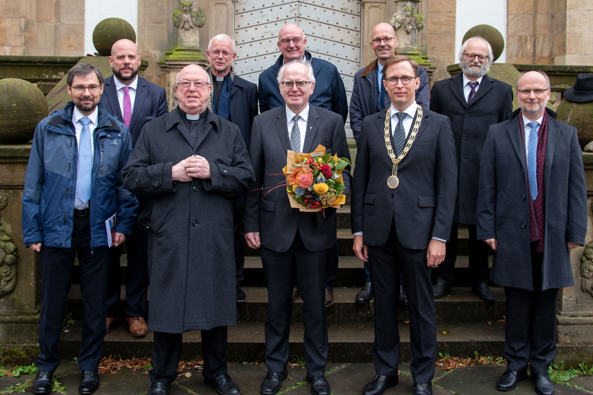 Abschied und Neuanfang: Emeritus Prof. Dr. Josef Meyer zu Schlochtern (Mitte) und die beiden designierten Lehrstuhlinhaber, PD Dr. Aaron Langenfeld (2. Reihe links) und Dr. Daniel Lanzinger (1. Reihe links) stehen gemeinsam mit dem Magnus Cancellarius, Erzbischof Hans-Josef Becker (2.v.l.), Rektor Prof. Dr. Stefan Kopp (4.v.l.) und den Professorenkollegen Andreas Koritensky (5.v.l.), Peter Schallenberg, Wolfgang Thönissen, Bernd Irlenborn und Ansgar Moenikes (2. Reihe v.l.) auf den Stufen zur Universität- und Marktkirche.