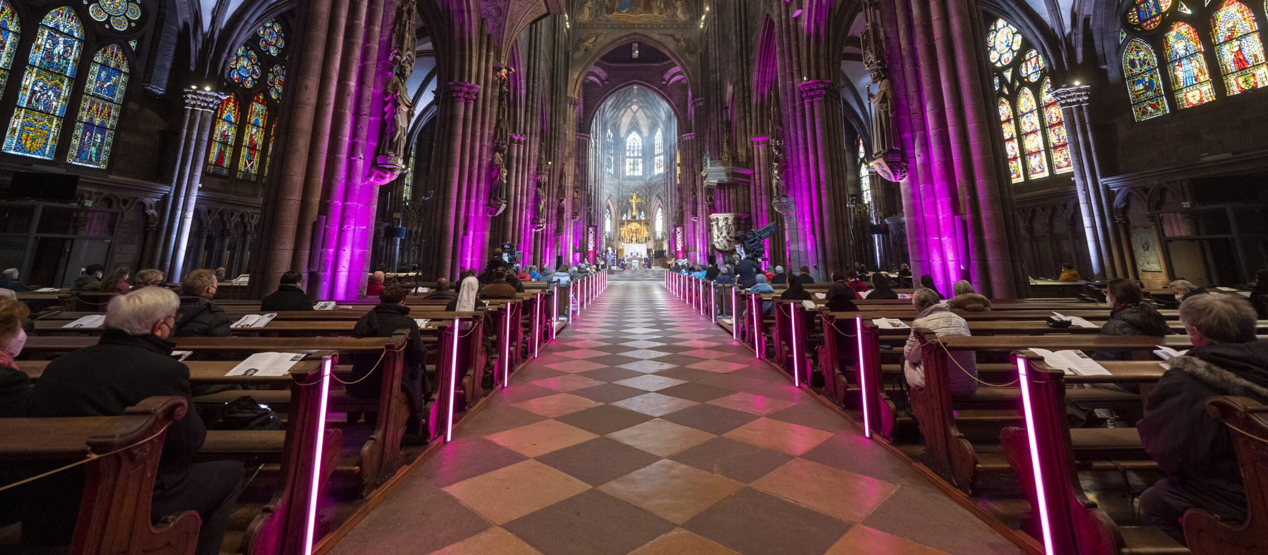 Viele Menschen in einer Kirche, lila Beleuchtung, Goldenes Kreuz im Hintergrund