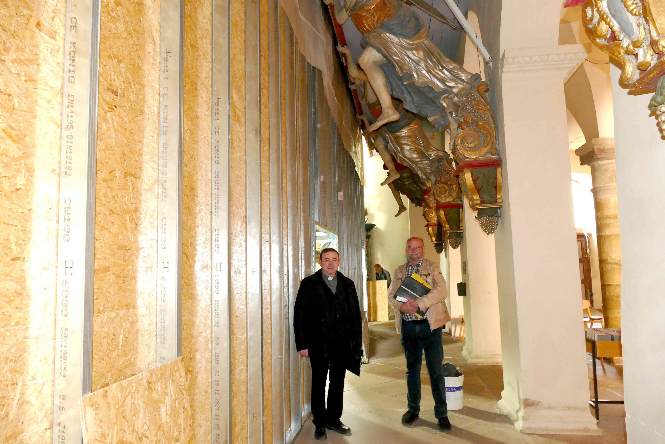 Die großen Engelatlanten, die die Orgelbühne tragen, schauen während der Bauarbeiten vor die Staubschutzwand. Sie werden zum Schutz noch eingepackt, kündigen Pfarrdechant Dr. Hans-Bernd Krismanek (links) und Architekt Albert Henne an.