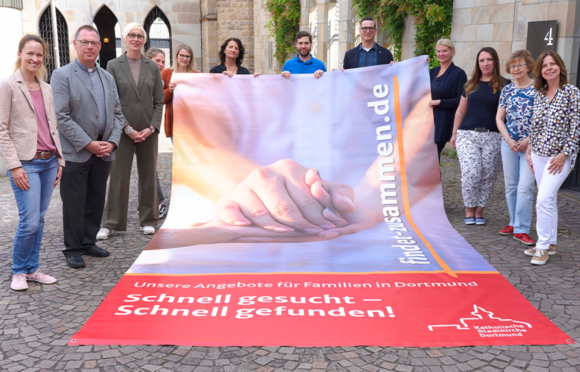 220621_pm_findet_zusammen: Mitglieder der katholischen Fachkonferenz Familie und der stellvertretende Stadtdechant Michael Ortwald (2.v.l.) entrollten im Propsteihof ein Banner zum neuen Internetangebot www.findet-zusammen.de. Foto: Michael Bodin / Kath. Pressestelle Dortmund