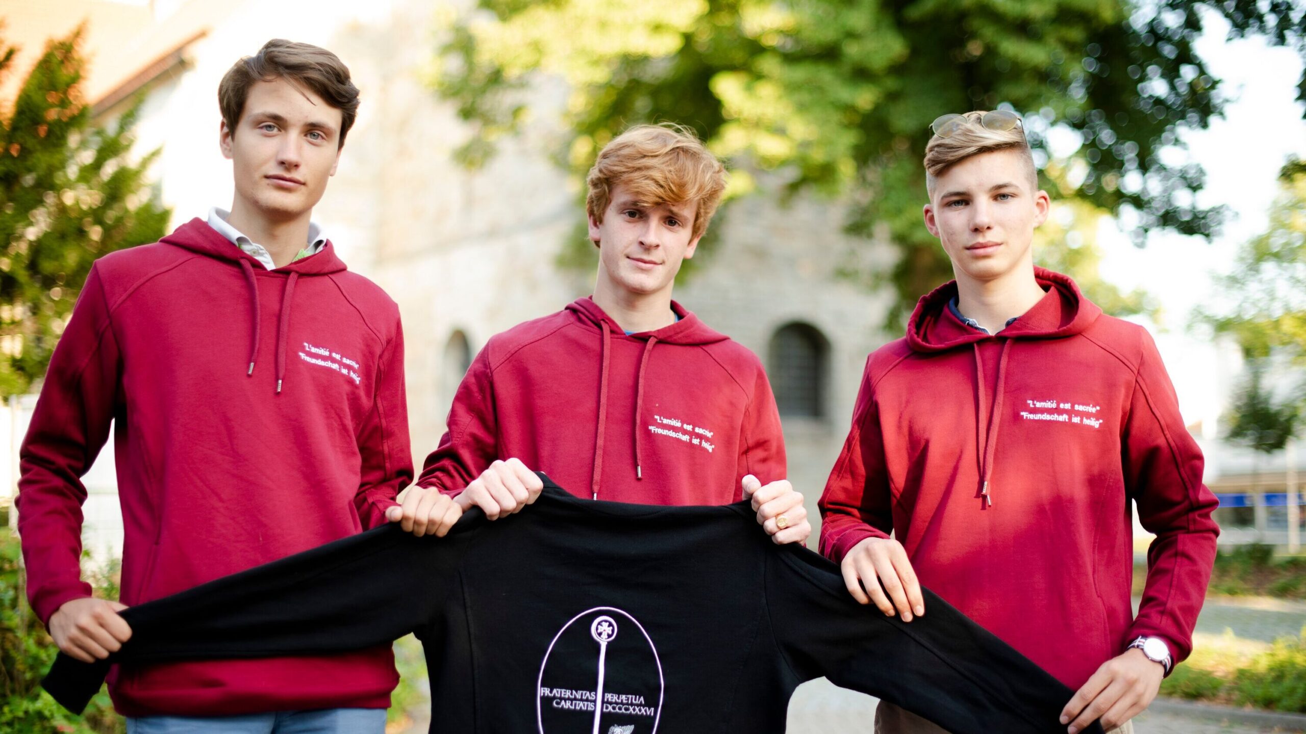 Drei Mitgleider der Jugendorganisation der Fraternität St. Liborius halten einen Pullover mit ihrem Logo darauf