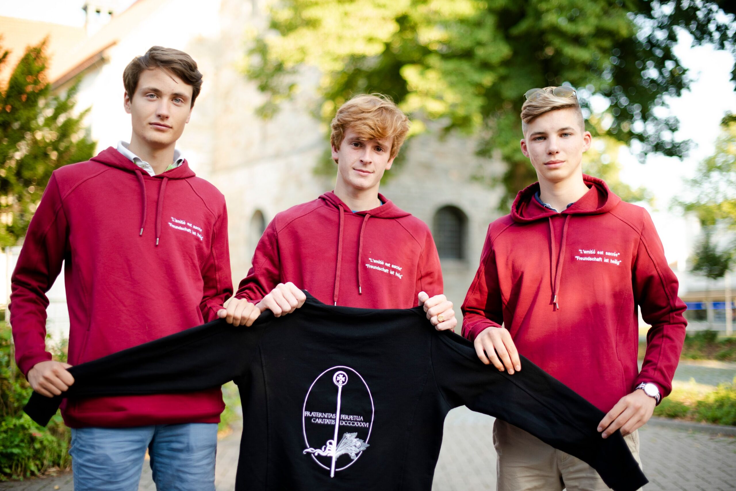 Drei Mitgleider der Jugendorganisation der Fraternität St. Liborius halten einen Pullover mit ihrem Logo darauf