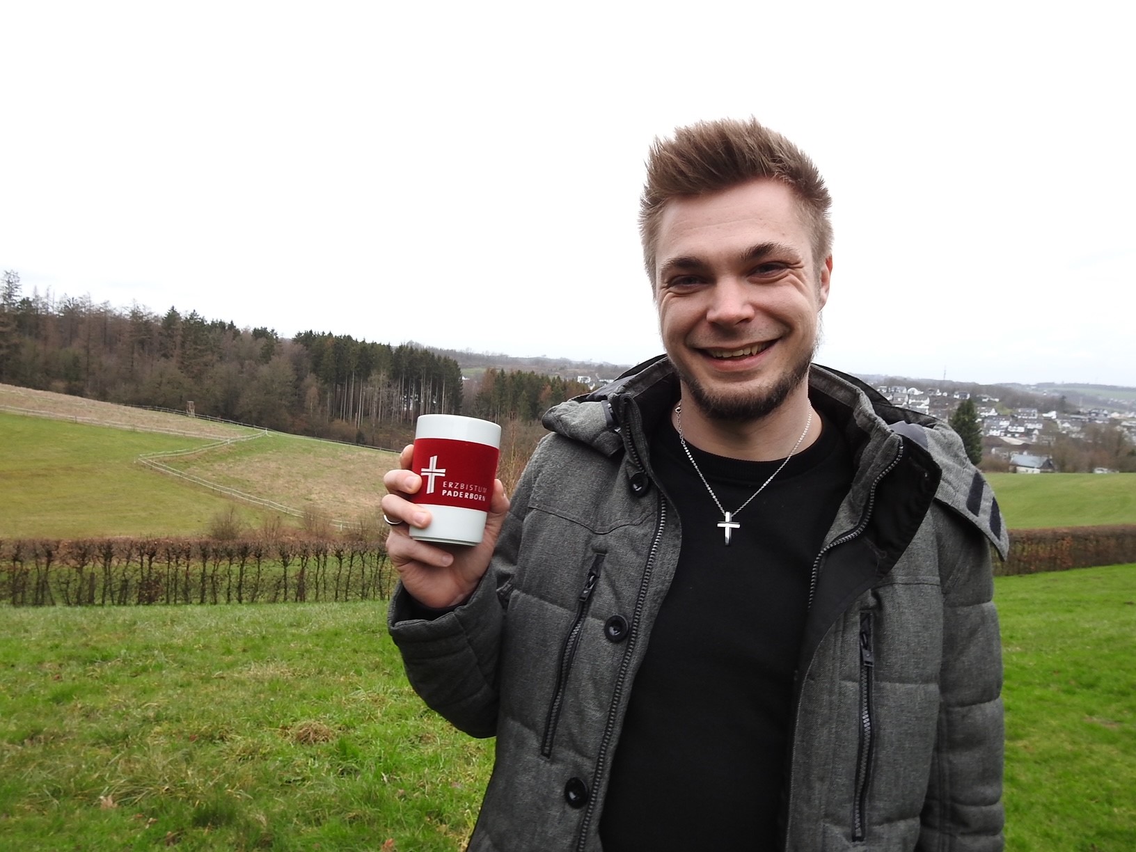 Robin Geisweid steht vor einem Feld. In der Hand hält er einen Kaffeebecher des Erzbistums Paderborn