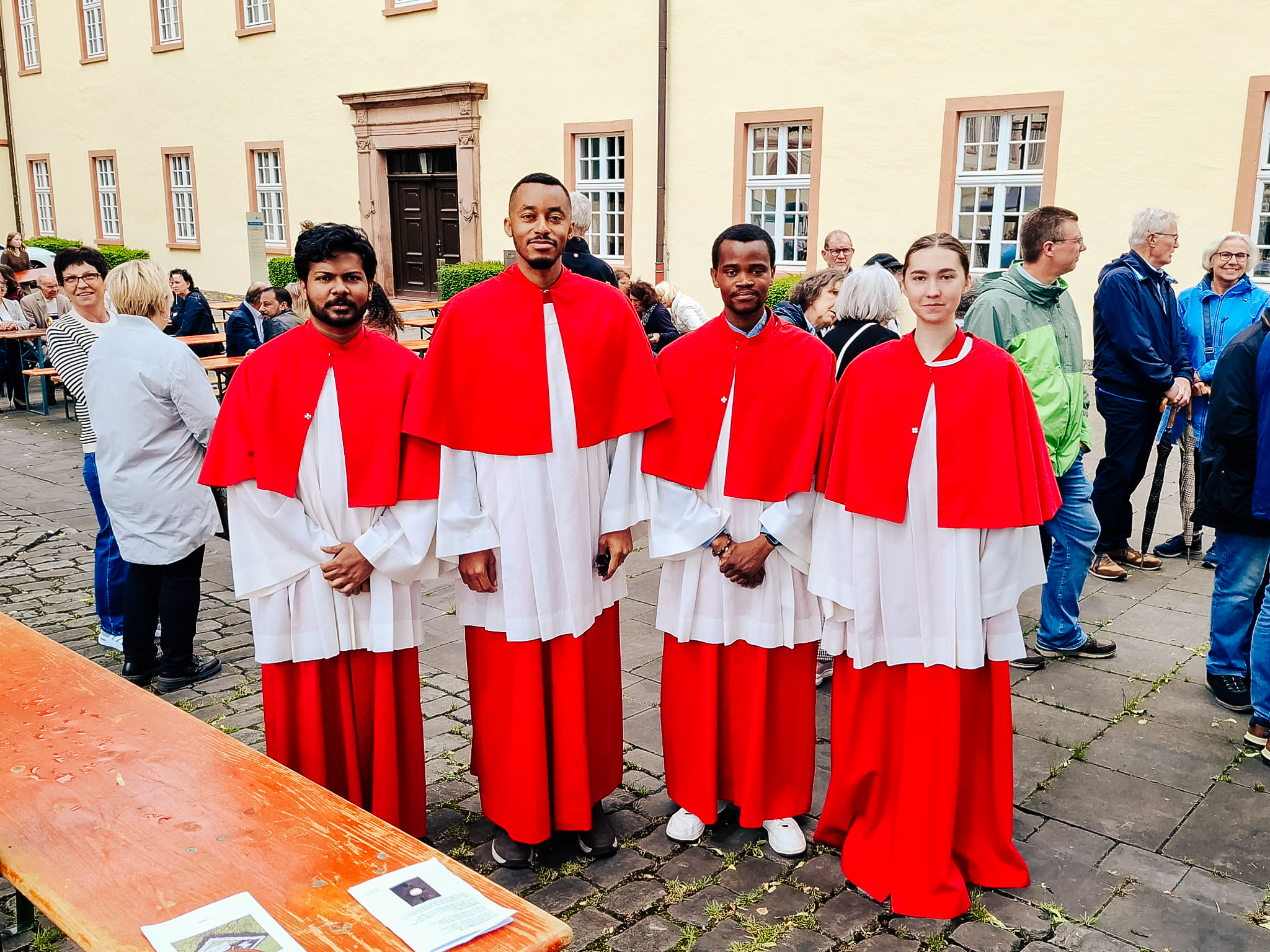 Vier Mitglieder von Come and see stehen in Messdienergewändern vor einer Kirche.