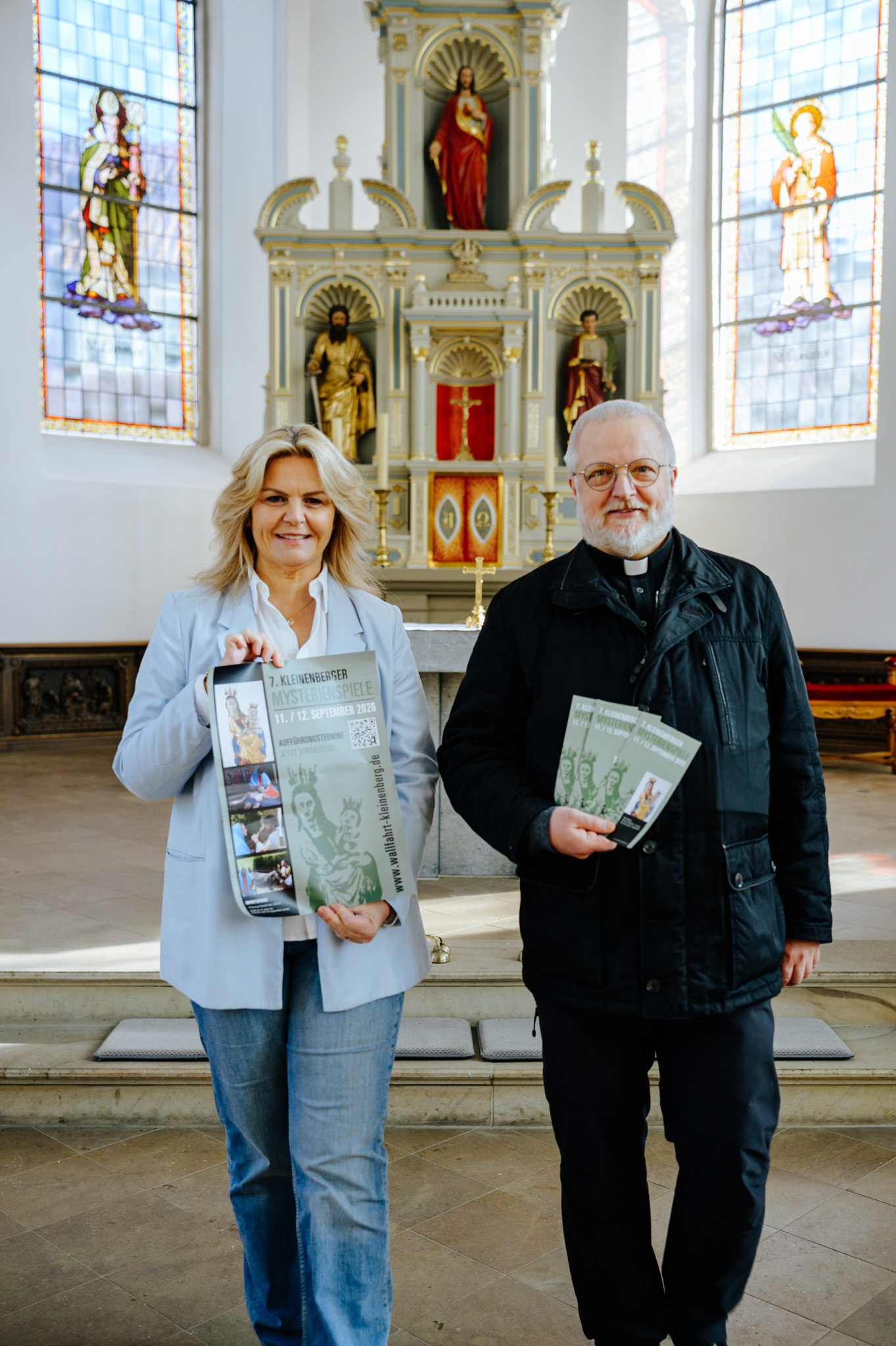 Nina-Maria Mehring und Pastor Stefan Stratmann in der Pfarrkirche St. Cyriakus mit Plakaten und Flyern für die Mysterienspiele in Kleinenberg.
