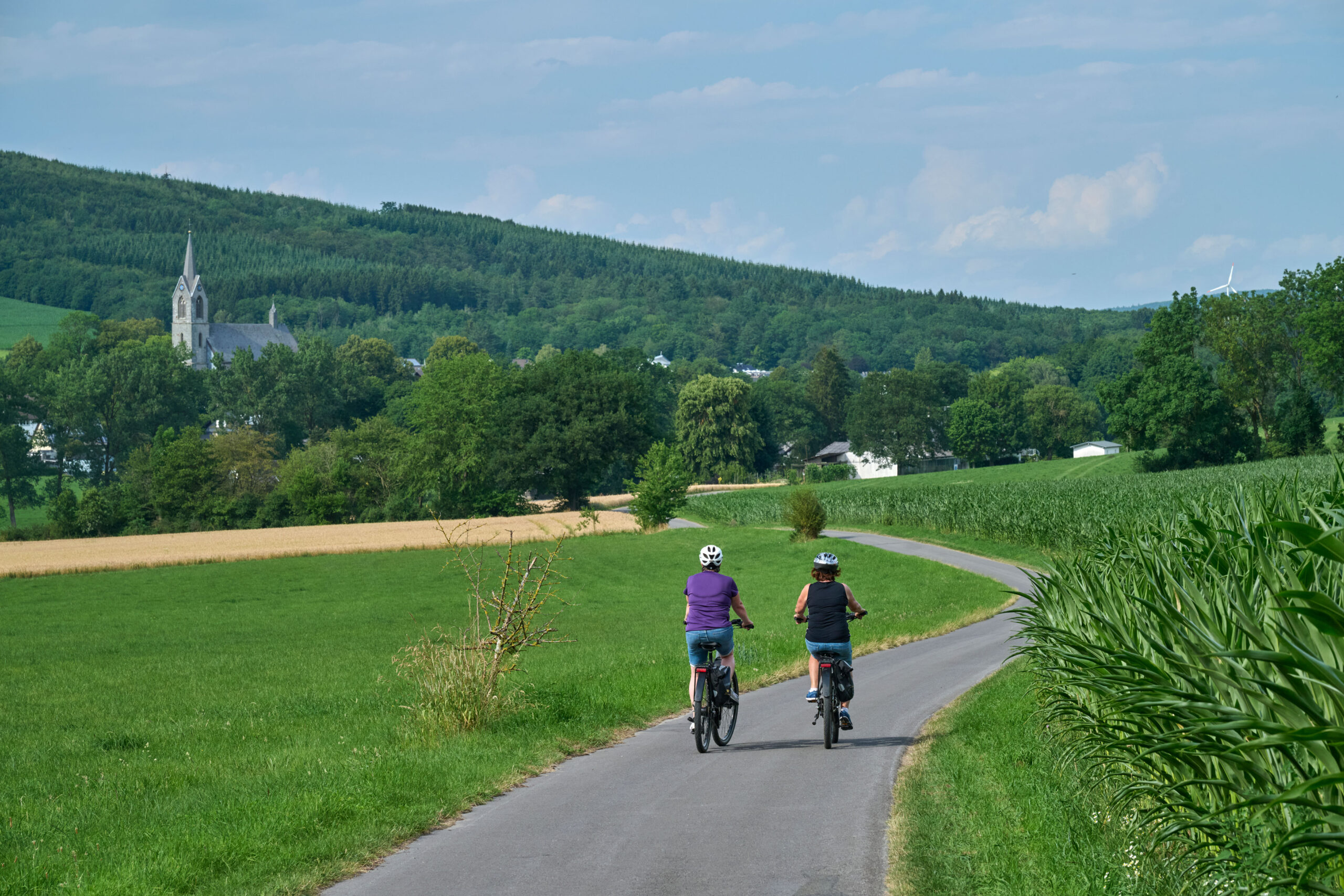Zwei Radfahrerinnen auf einer Straße durch grüne Felder, im Hintergrund St. Pankratius in Reiste.
