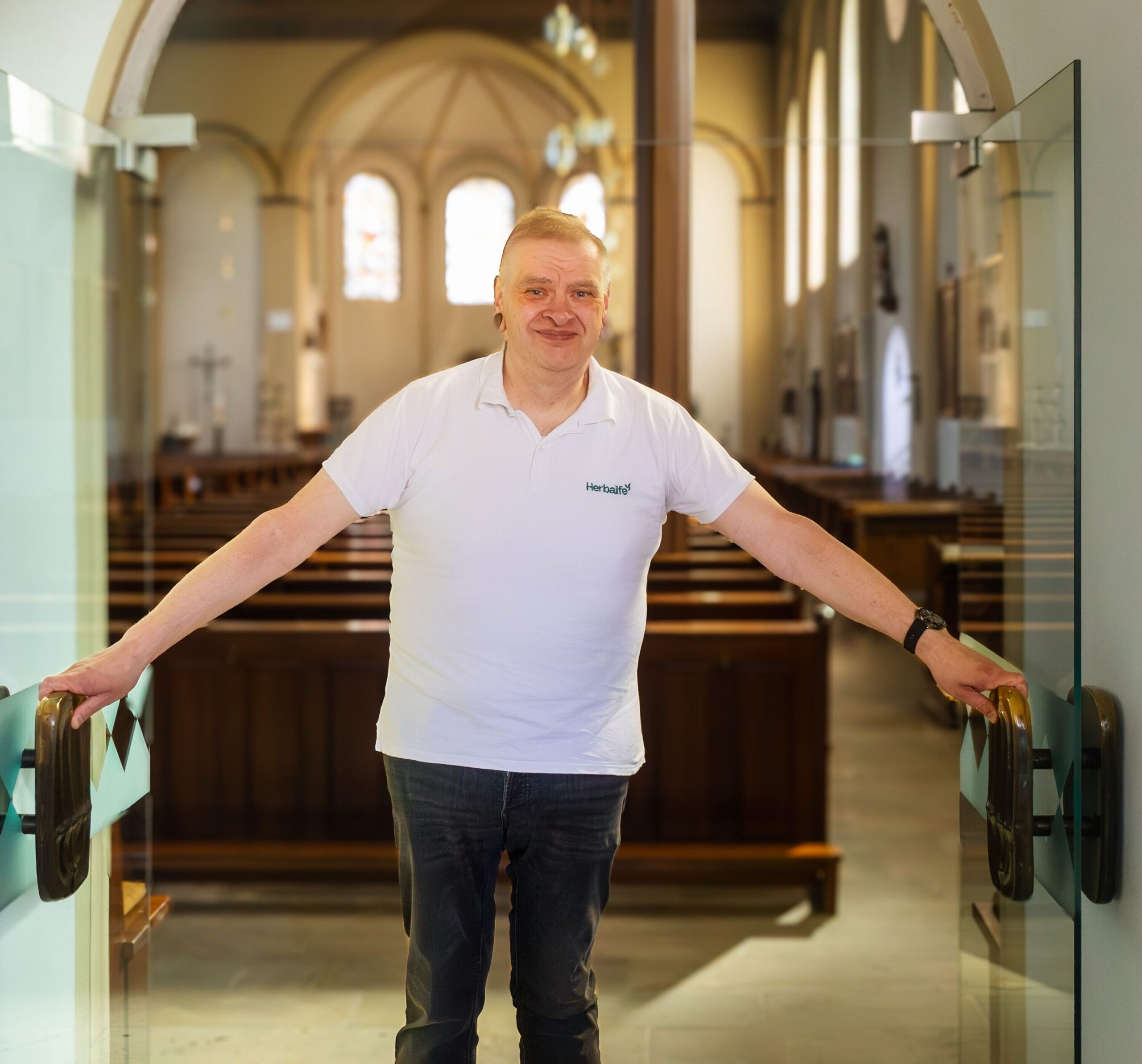 Ein Mann in weißem T-Short öfnet die Tür zu einer Kirche und lächelt in die Kamera