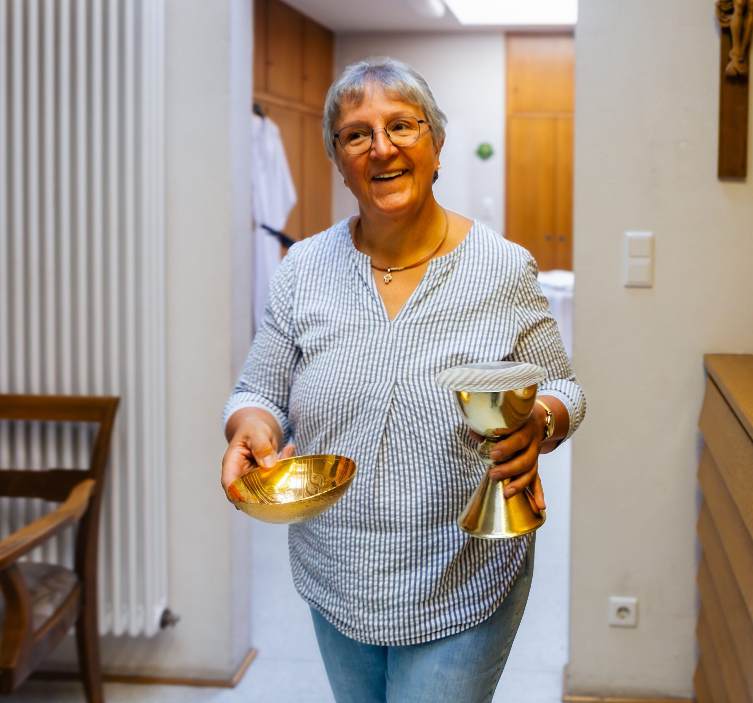 Aufnahme einer älteren Frau in Bluse, die eine goldene Schale und einen goldenen Kelch in der Hand hält.
