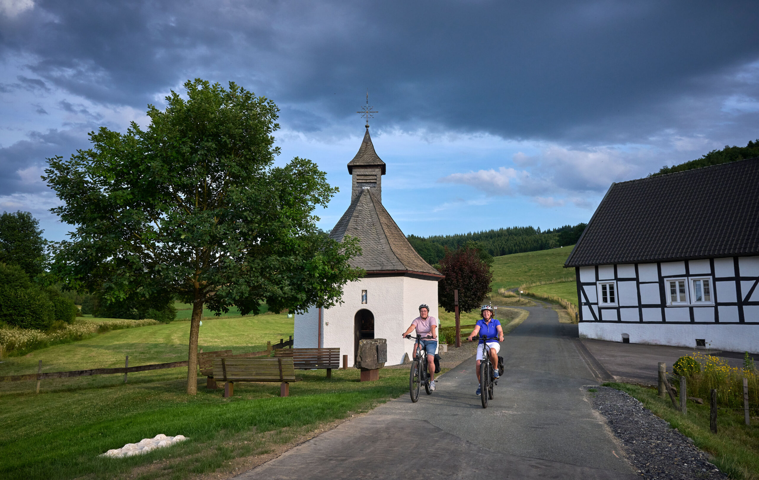 Zwei Radfahrerinnen fahren an St. Jakobus in Bremscheid und einem Fachwerkhaus vorbei.