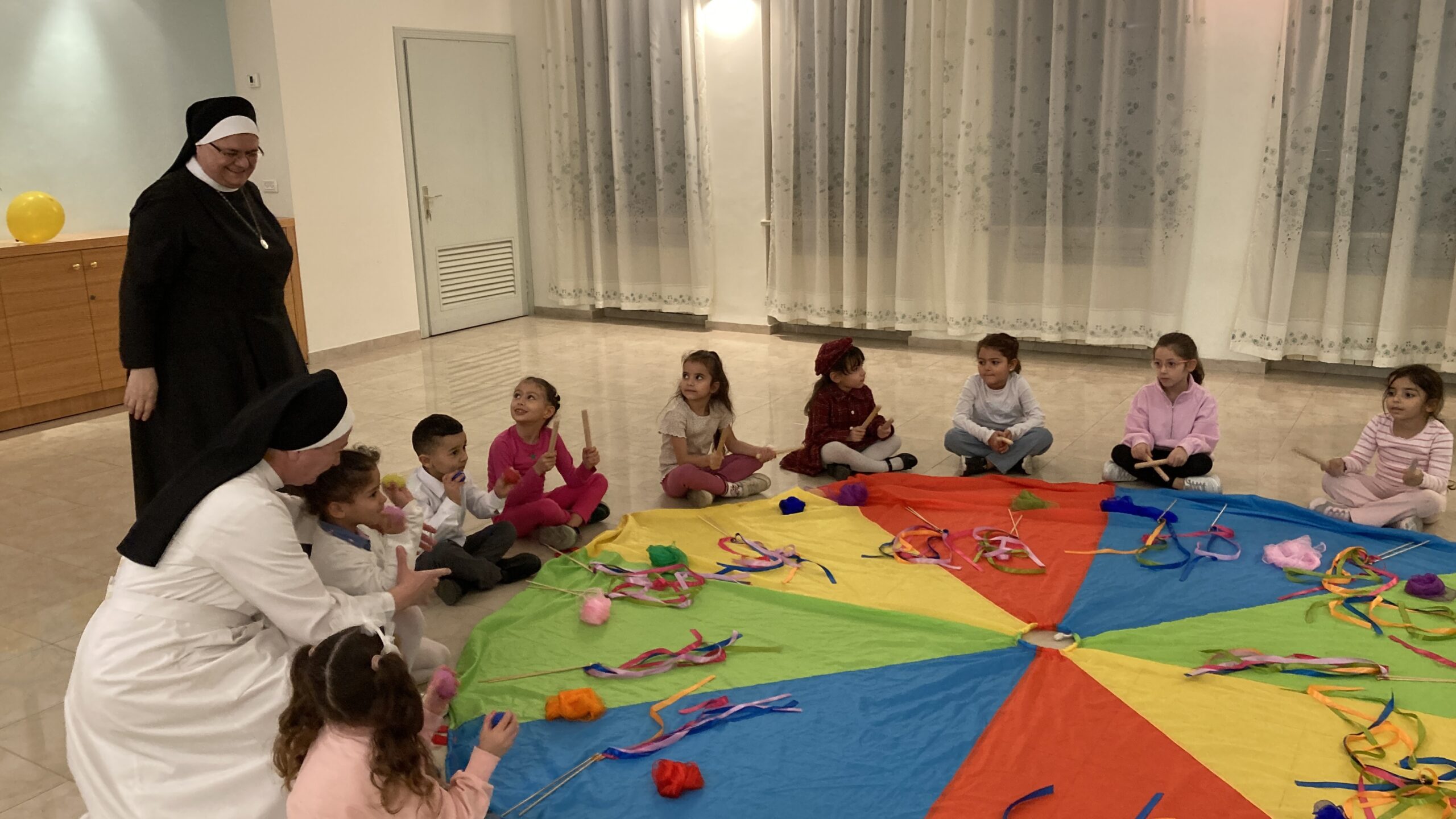 Zwei Ordensschwestern begleiten mehrere Kinder, die im Kreis um ein großes, buntes Tuch sitzen und mit Holzstäben spielen.