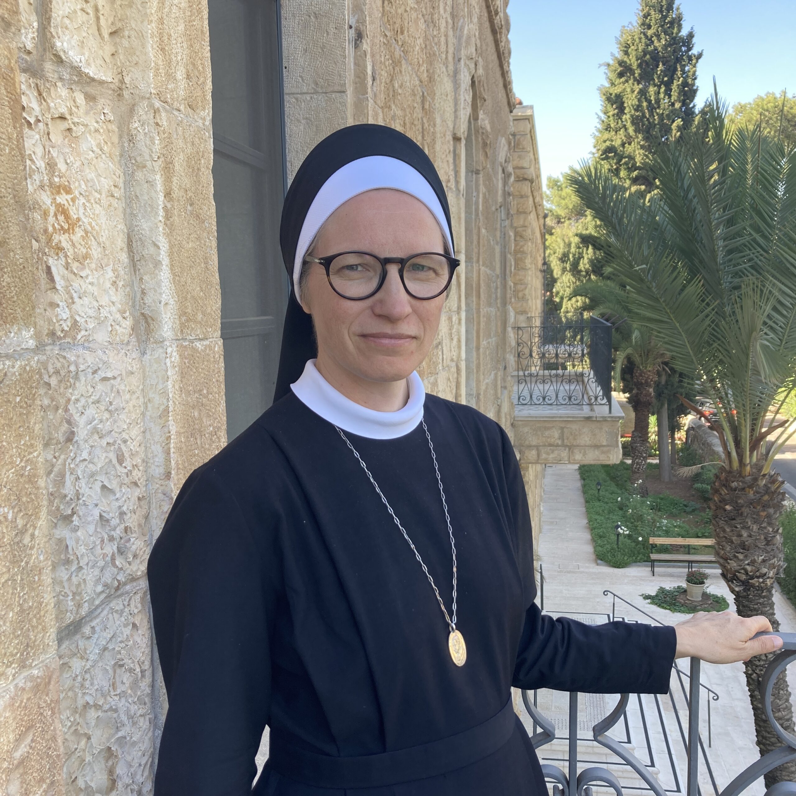 Eine Ordensschwester mit Brille, schwarzem Habit und weißem Kragen steht vor einer hellen Steinmauer auf einem Balkon. Im Hintergrund sind Palmen und weitere Bäume zu sehen.